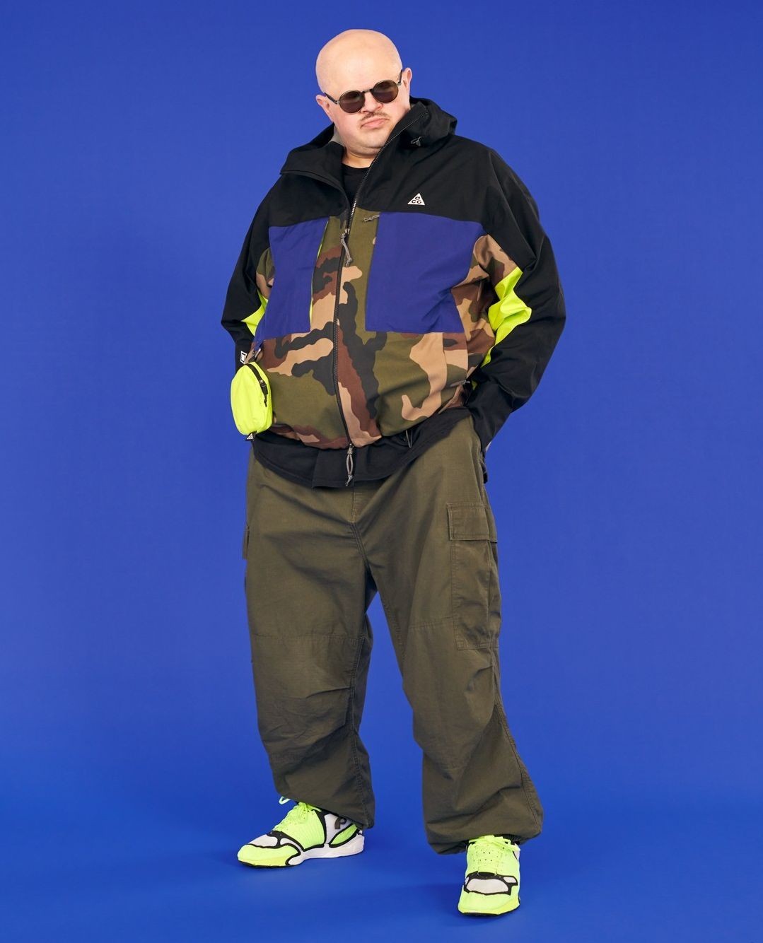 Full-length shot of a bald man with sunglasses wearing a camouflage jacket, olive green pants, and neon yellow sneakers against a blue background.