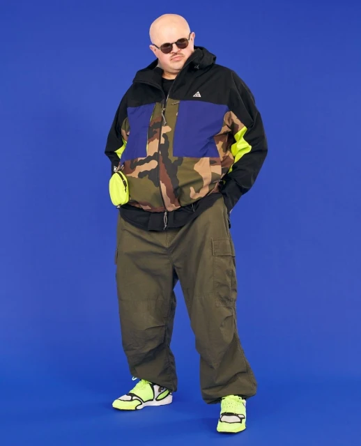 Full-length shot of a bald man with sunglasses wearing a camouflage jacket, olive green pants, and neon yellow sneakers against a blue background.