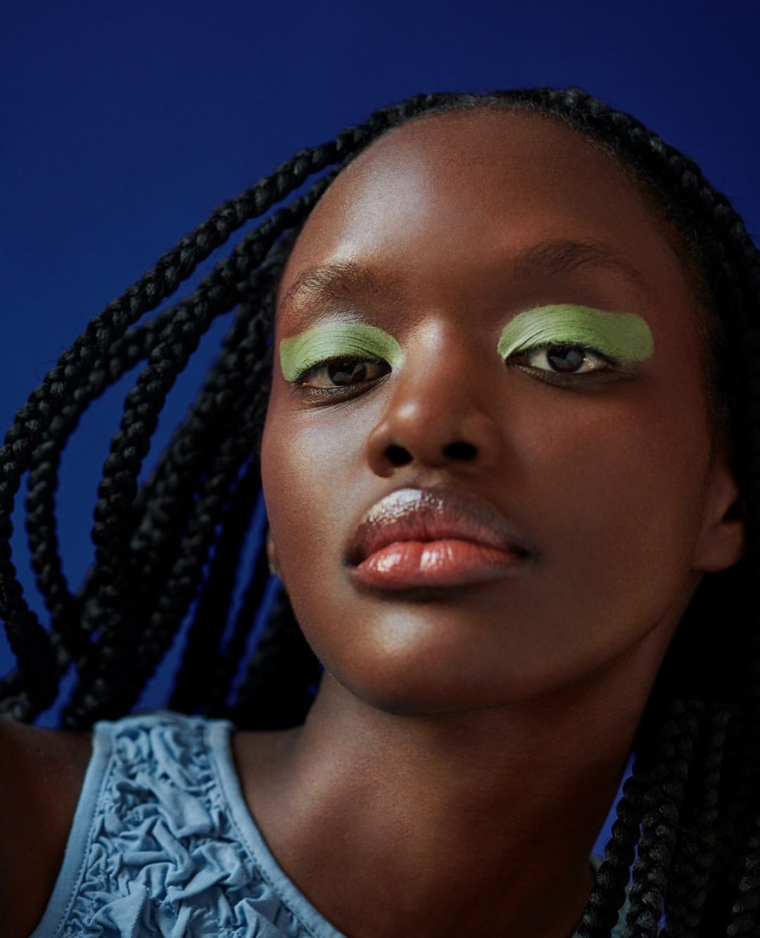 Close-up portrait of an adult female model with dark skin and braided hair. She wears green eye shadow and a blue ruffled top against a blue backdrop.