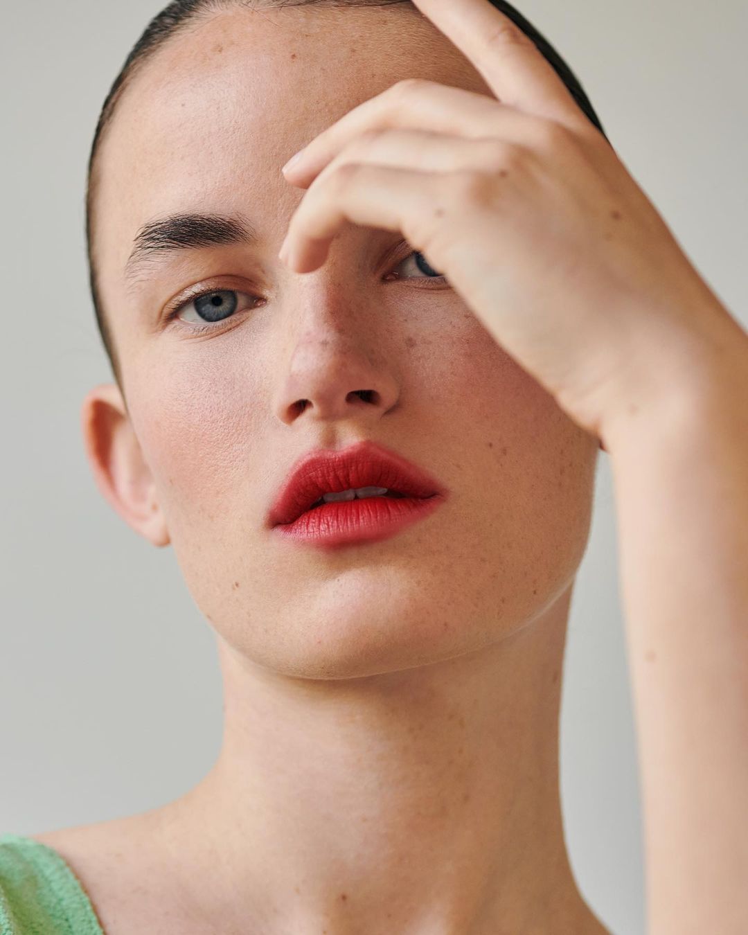 Close-up portrait of a woman with red lips and blue eyes, hand raised near her forehead against a plain background.