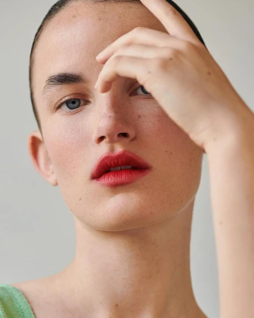Close-up portrait of a woman with red lips and blue eyes, hand raised near her forehead against a plain background.