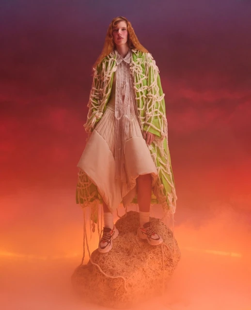 Fashionable woman in an avant-garde outfit stands confidently on a rock formation against a dramatic, colorful sky backdrop.