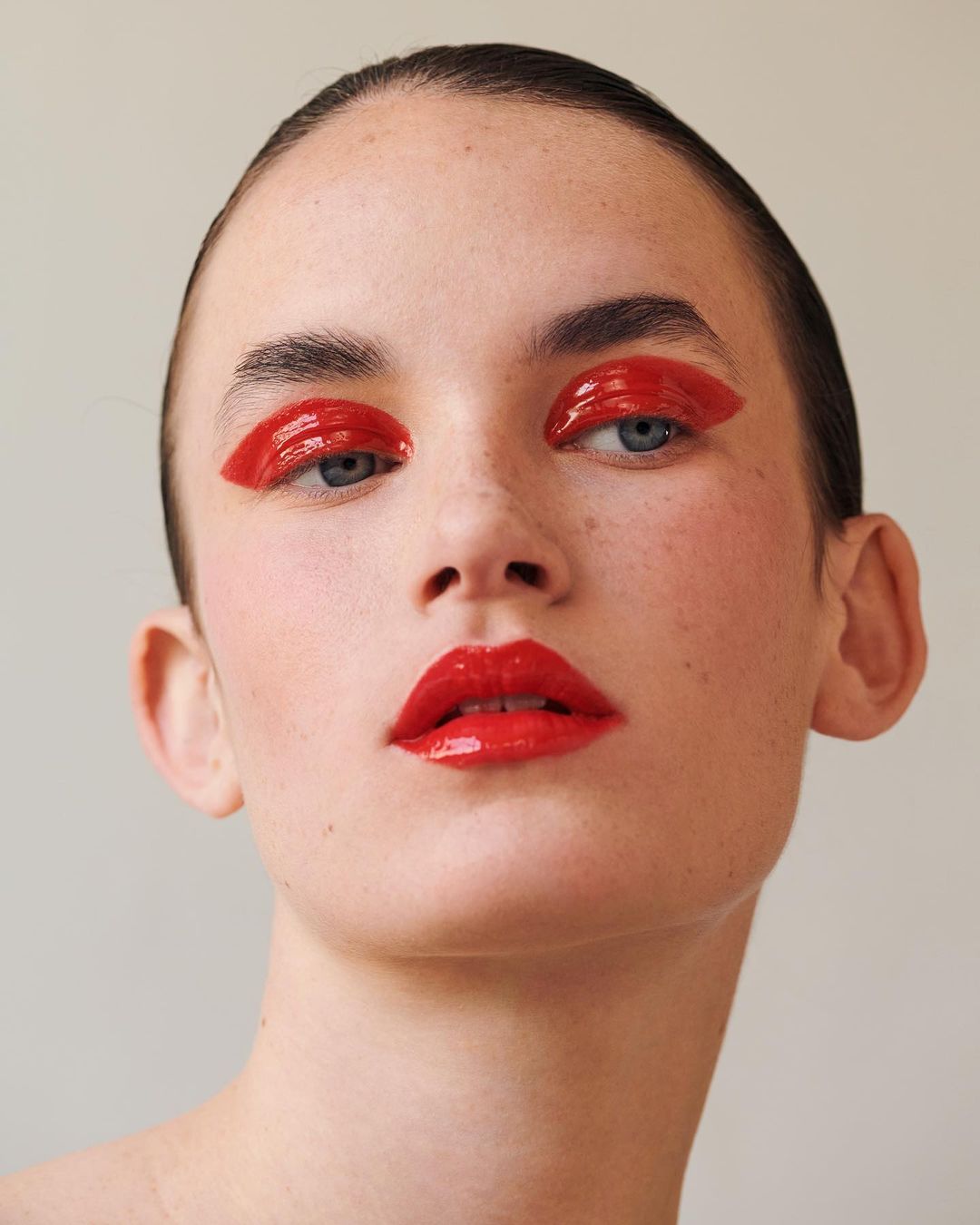 Close-up portrait of a young woman with red glossy makeup on her eyes and lips against a neutral background in a beauty shot.
