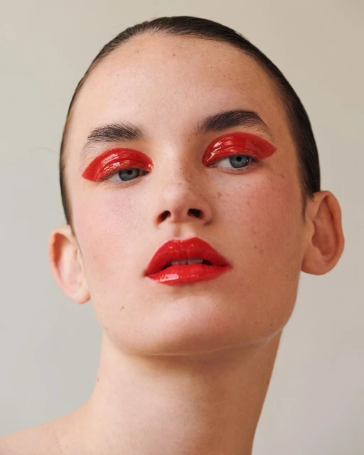 Close-up portrait of a young woman with red glossy makeup on her eyes and lips against a neutral background in a beauty shot.