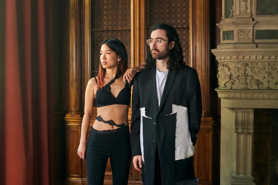 Indoor fashion shot of an Asian woman and Caucasian man in black outfits posing by a decorative fireplace and wood-panelled wall.
