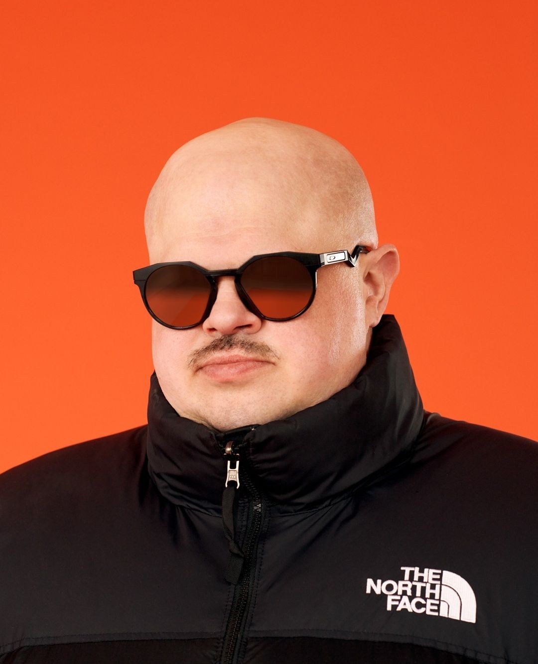 Portrait of a bald man wearing sunglasses and a black puffer jacket against an orange backdrop in a studio setting.