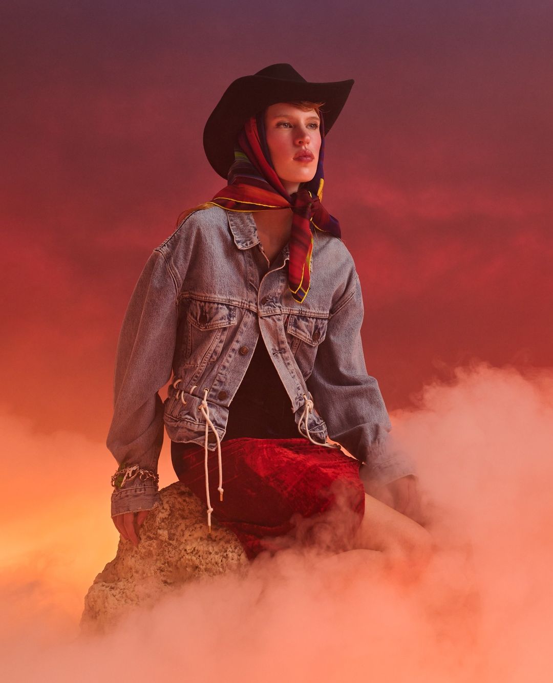 Woman in cowboy hat and denim jacket sits on a rock amid fog, illuminated by warm light with a red and purple gradient background.
