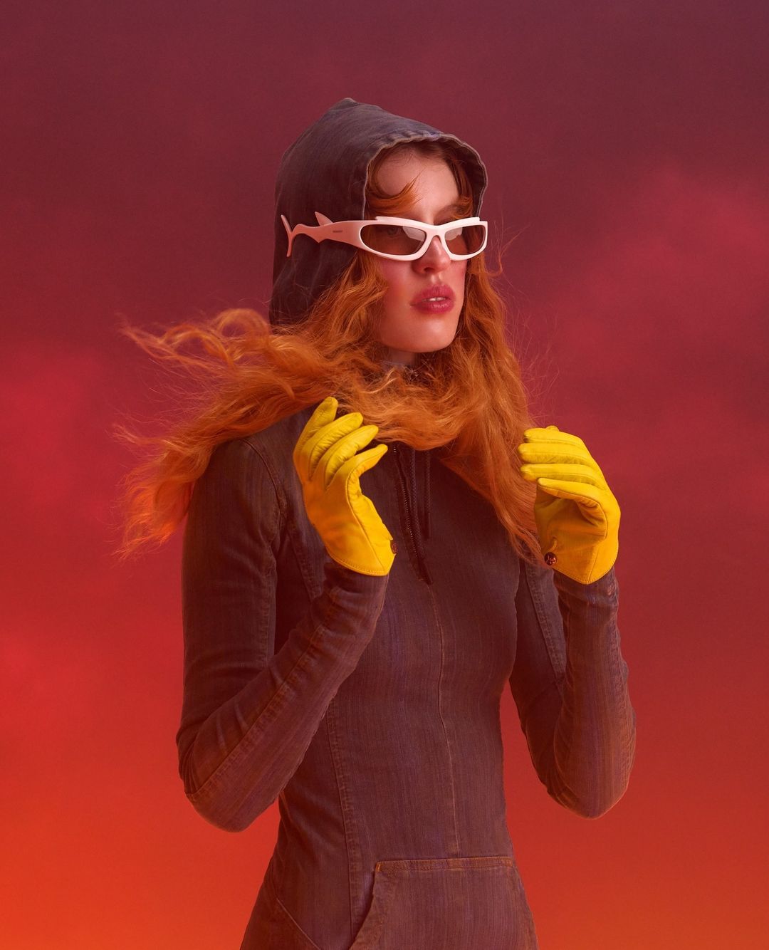 A person with auburn hair wearing sunglasses and yellow gloves poses against a red gradient background in a hooded, form-fitting dress.