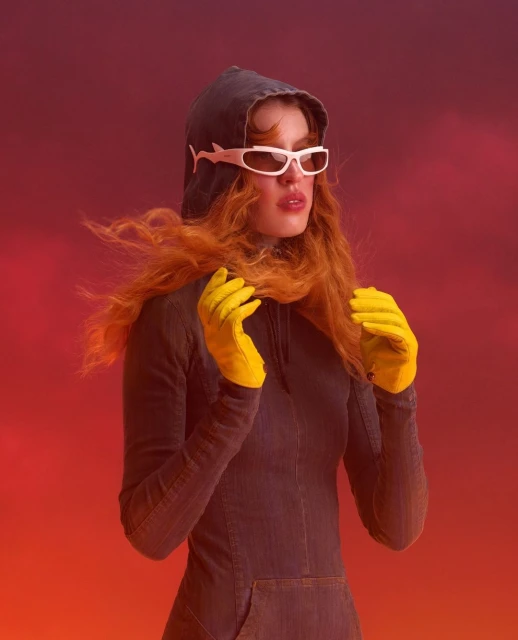 A person with auburn hair wearing sunglasses and yellow gloves poses against a red gradient background in a hooded, form-fitting dress.