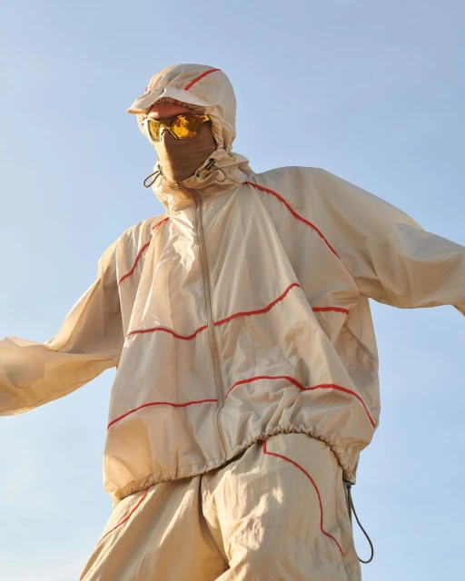 Person wearing a hooded, transparent tracksuit with orange piping, sunglasses, and a face covering against a light blue sky.