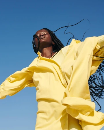 Black woman with braided hair wearing a yellow shirt against a clear blue sky, photographed from a low angle by Lucie Hugary.