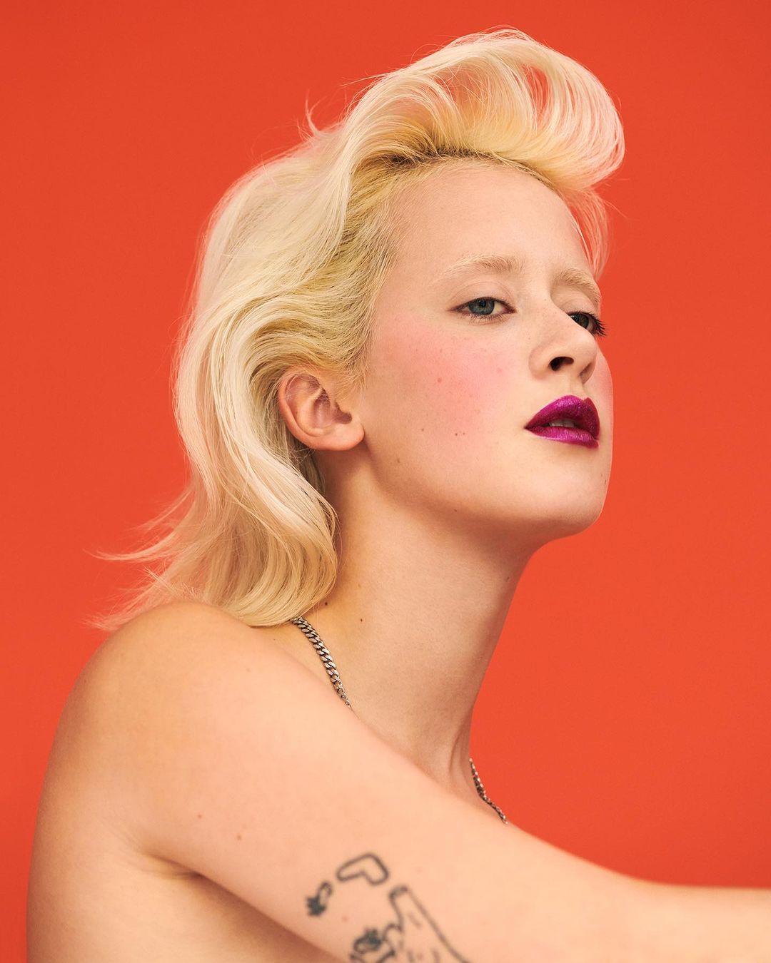 Portrait of a young adult woman with platinum blonde hair, a silver chain necklace, and a tattoo against an orange backdrop.