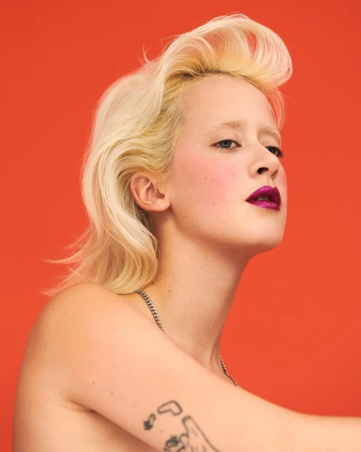 Portrait of a young adult woman with platinum blonde hair, a silver chain necklace, and a tattoo against an orange backdrop.