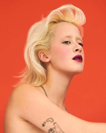Portrait of a young adult woman with platinum blonde hair, a silver chain necklace, and a tattoo against an orange backdrop.