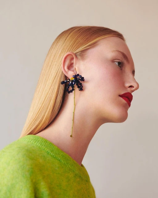 Profile of a blonde woman with red lipstick, wearing a bright green sweater and flower-shaped earrings with a long stem and dark blue beads.