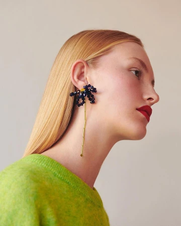 Profile of a blonde woman with red lipstick, wearing a bright green sweater and flower-shaped earrings with a long stem and dark blue beads.