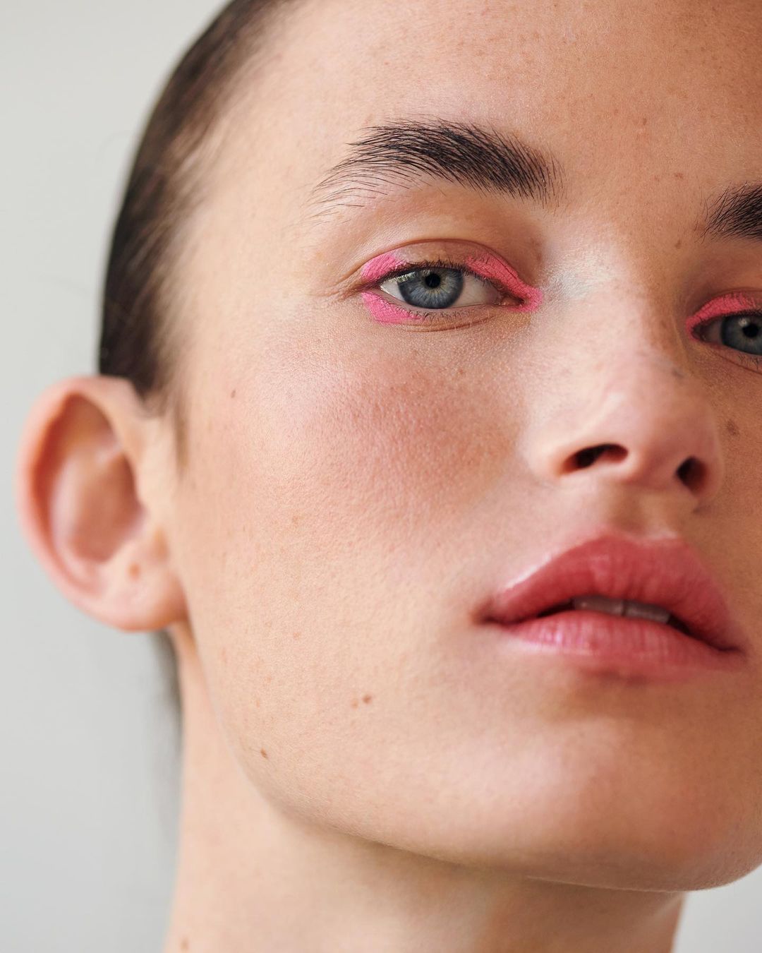 Close-up of a woman's face with light skin and pink eyeliner, focusing on the eyes, nose, and mouth in a beauty or fashion context.