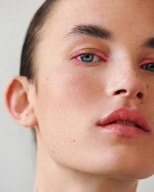 Close-up of a woman's face with light skin and pink eyeliner, focusing on the eyes, nose, and mouth in a beauty or fashion context.