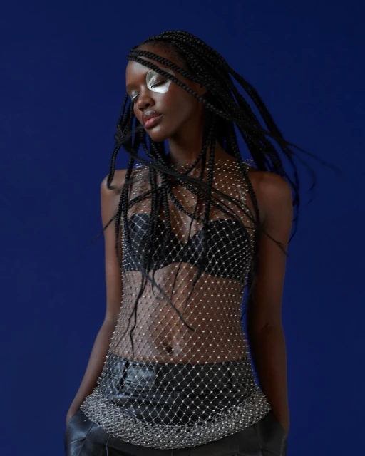 Young Black woman in a fishnet top with creative silver makeup posing against a dark blue background in a fashion shot.