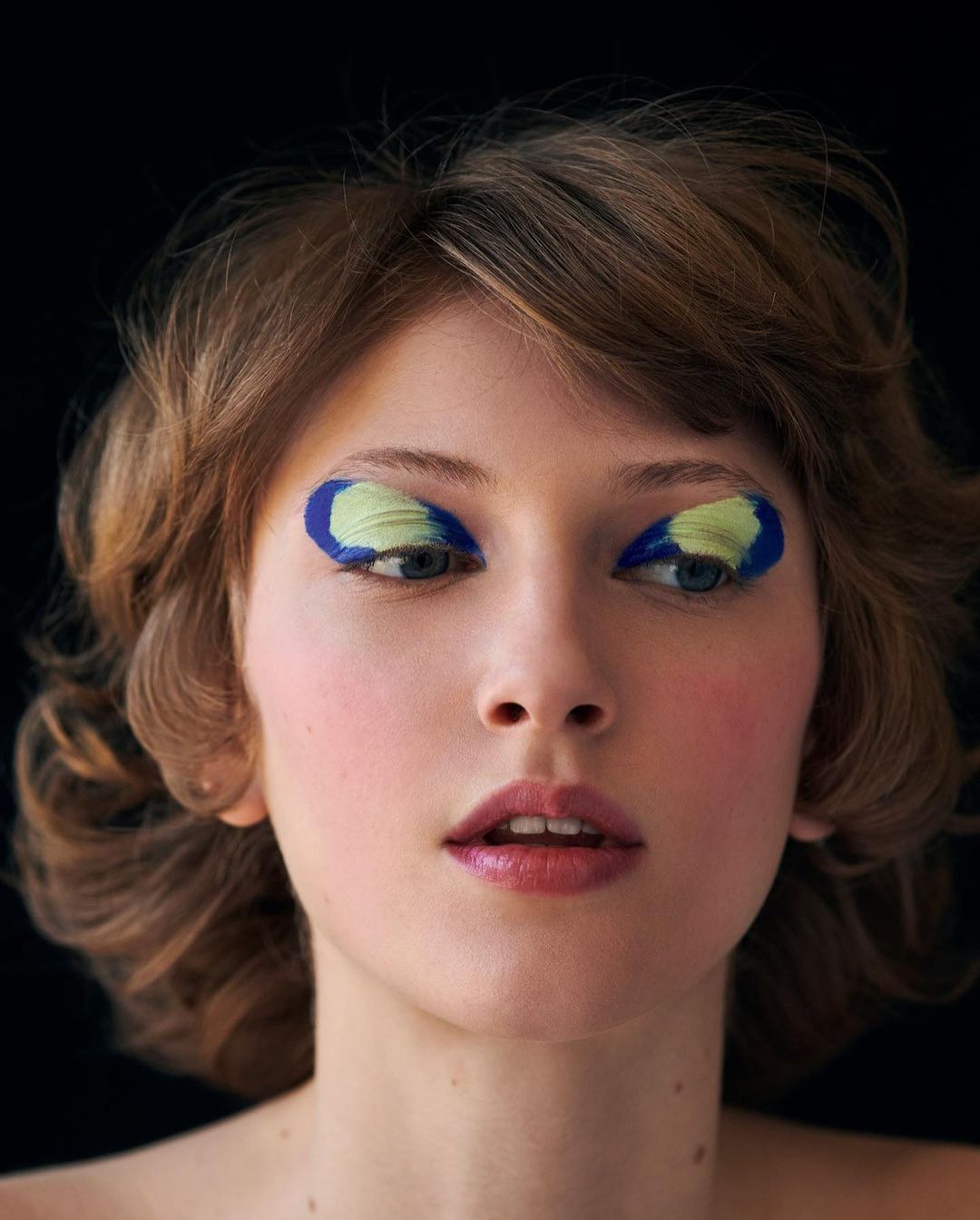Close-up of an adult female with short brown hair and dramatic blue and yellow eye makeup posing against a dark background.