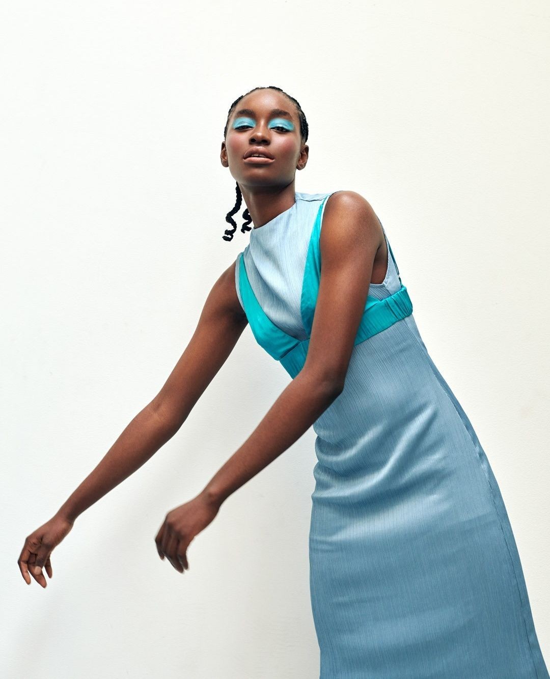 An African-American woman in a light blue dress with turquoise accents poses in front of a white wall, showcasing contemporary fashion.