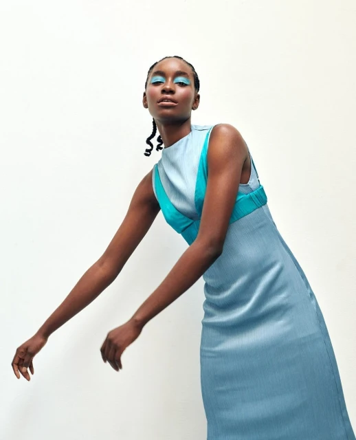 An African-American woman in a light blue dress with turquoise accents poses in front of a white wall, showcasing contemporary fashion.