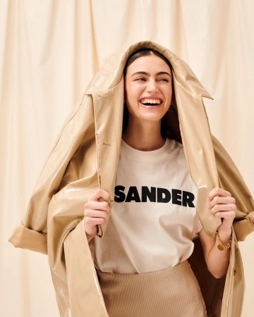 Smiling woman wearing a beige t-shirt holding a beige coat draped over her head against a pleated backdrop
