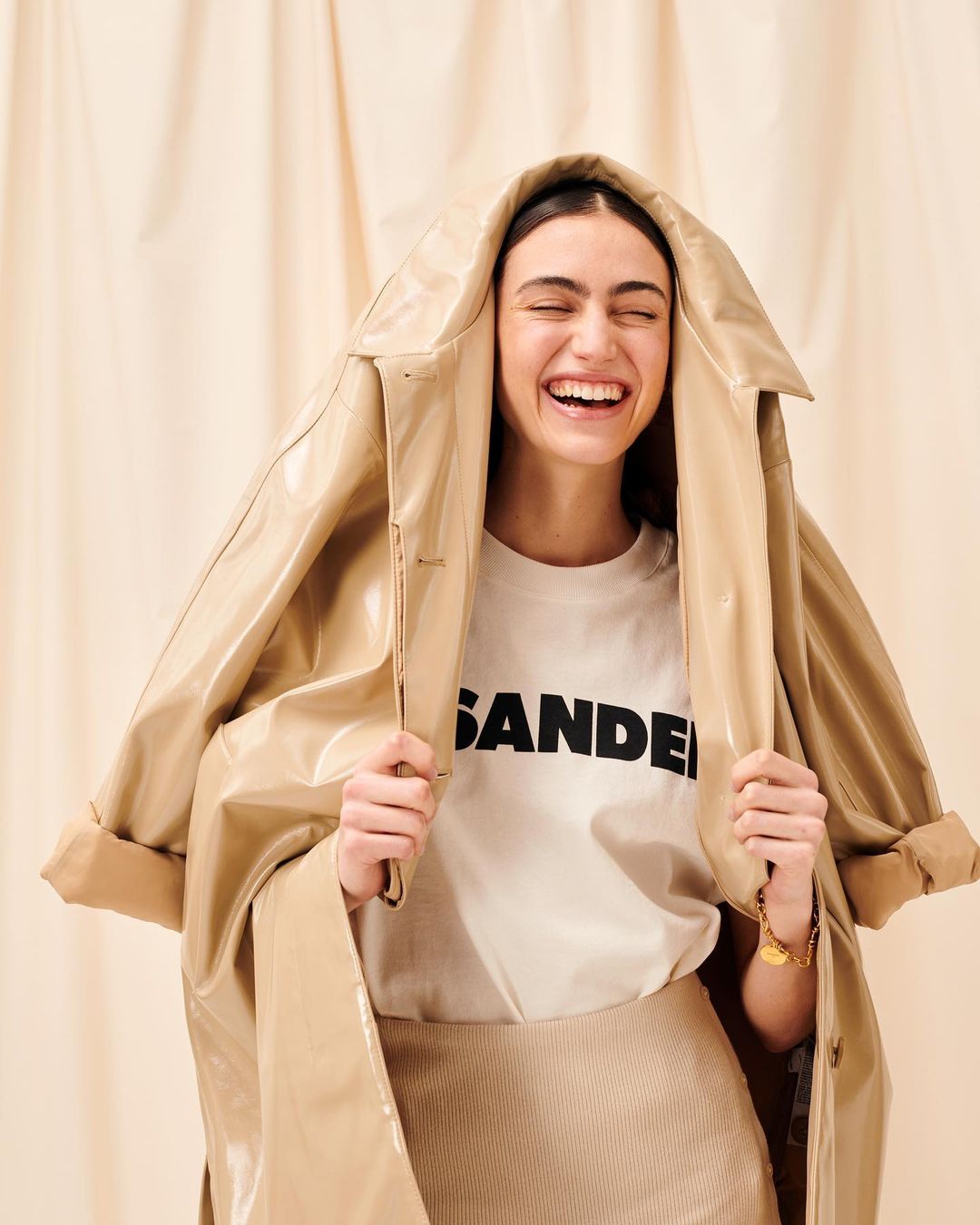 Smiling woman wearing a glossy beige coat draped over her head and shoulders, with a white t-shirt and skirt against a draped beige backdrop.