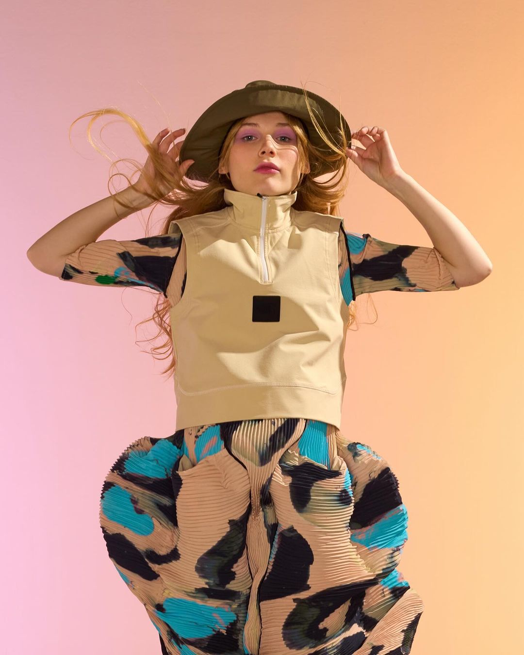 A young adult female model posing with hands on her hat, wearing a beige vest and patterned skirt, shot in a photo studio on a gradient background.
