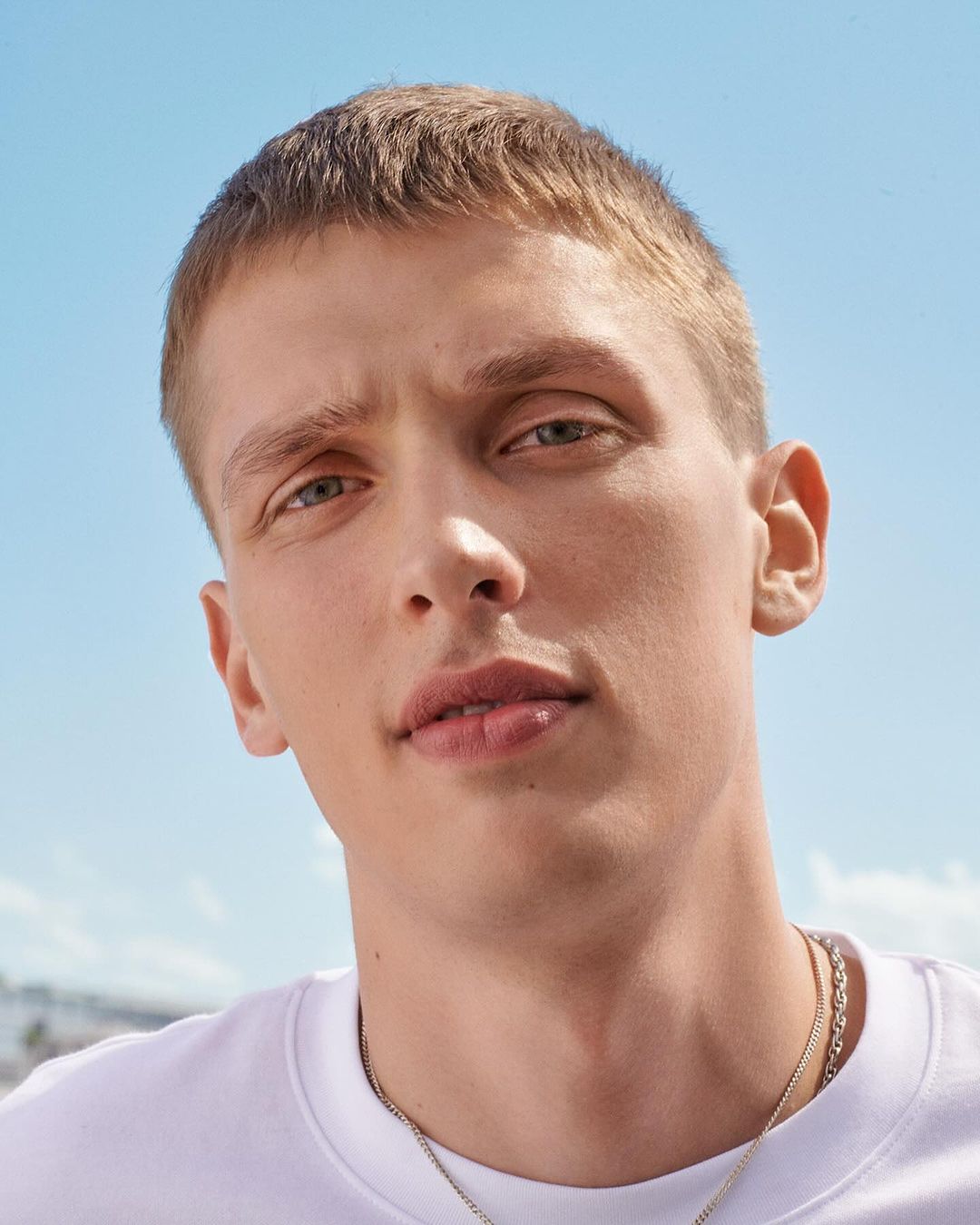 Close-up portrait of a young adult male with short blond hair, wearing a white t-shirt and silver chain against a light blue sky background.