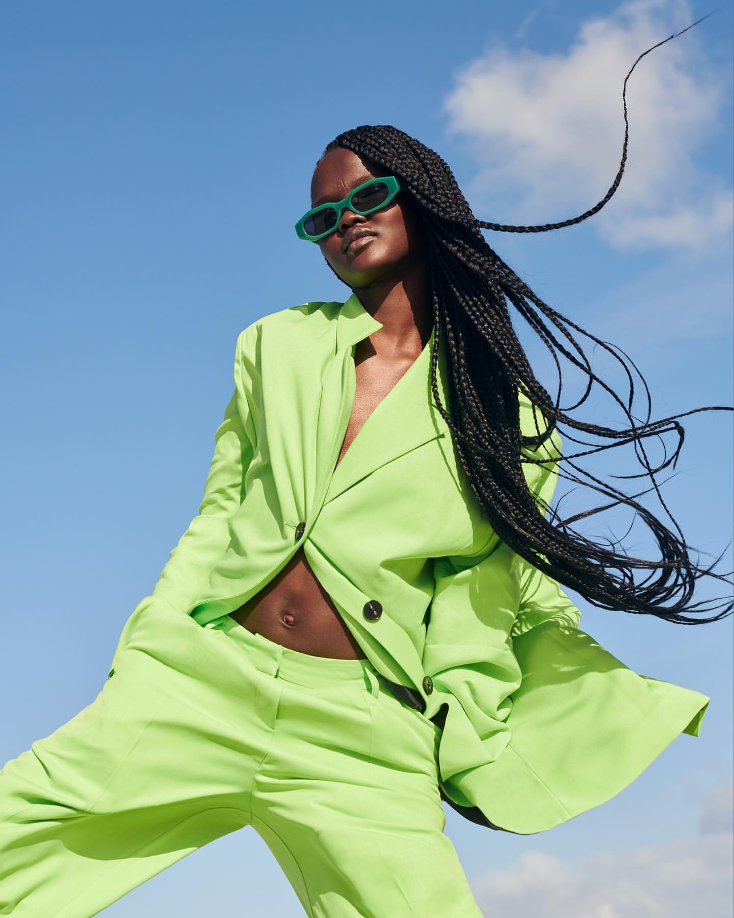 A confident Black woman in a lime-green suit strikes a dynamic pose against a blue sky backdrop, complemented by stylish sunglasses and braids.