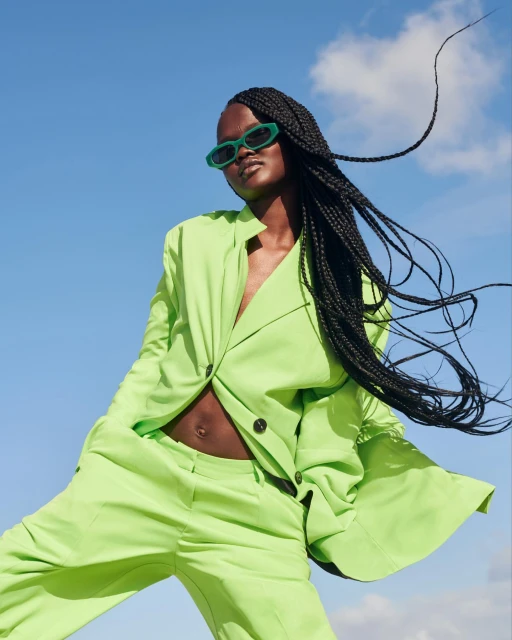 A confident Black woman in a lime-green suit strikes a dynamic pose against a blue sky backdrop, complemented by stylish sunglasses and braids.