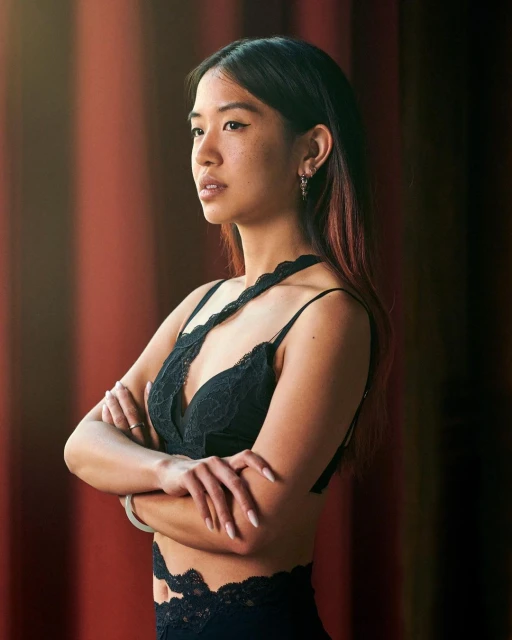 Portrait of a young Asian woman with long dark hair wearing a black lace top, standing with arms crossed in front of a red curtain.