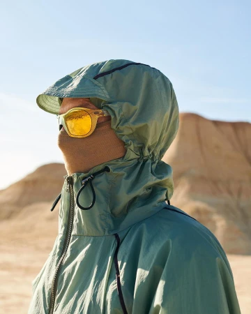 Person wearing yellow sunglasses and a green hooded jacket stands in a desert landscape under a clear sky looking into the distance.
