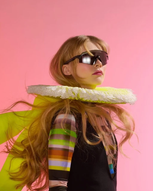 A woman with long blonde hair and dark sunglasses poses against a pink backdrop, wearing a black top and a wide, frilly white collar.