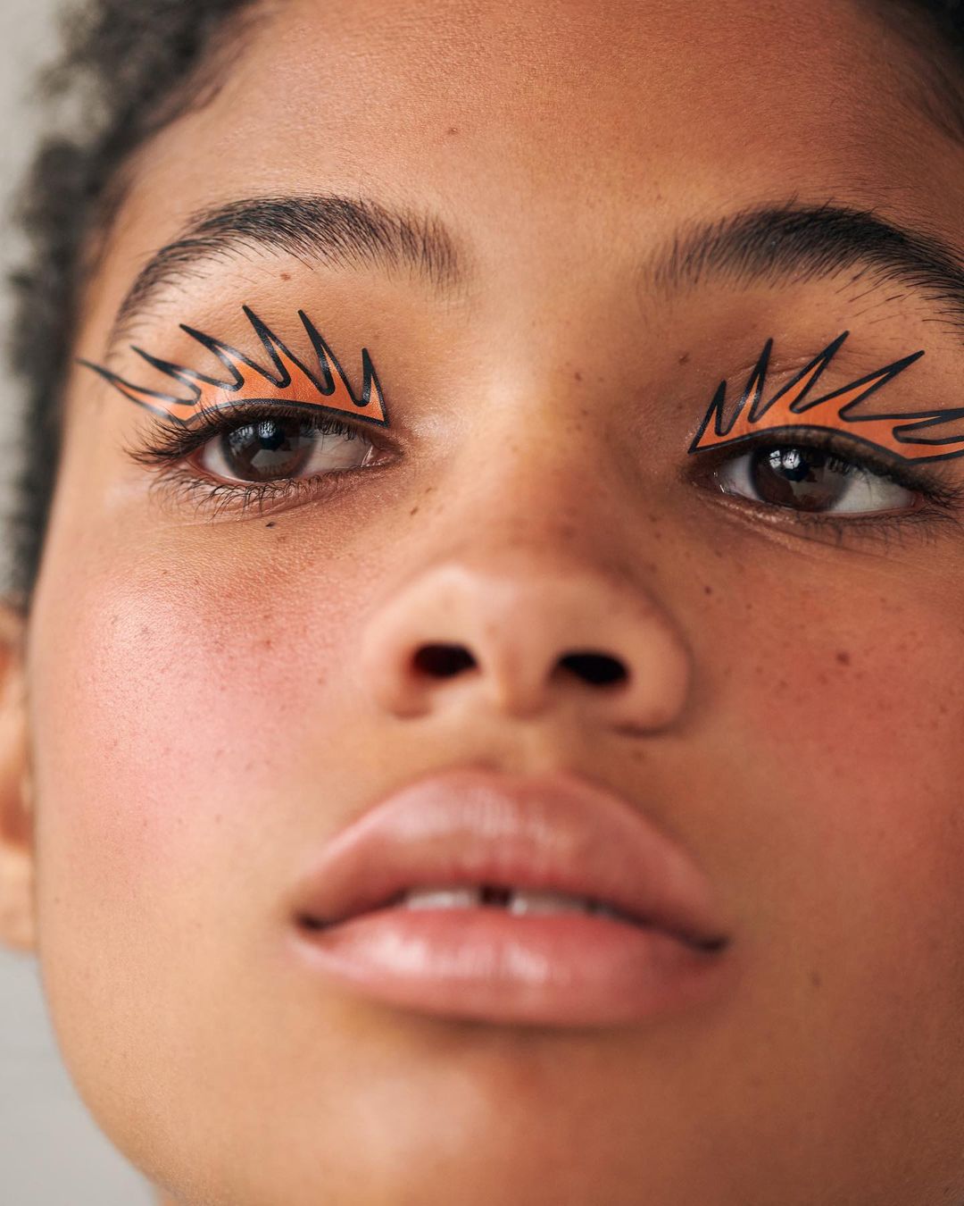 Close-up studio shot of a person with creative makeup featuring graphic orange eyeliner wings, neutral lip gloss, and fair skin.