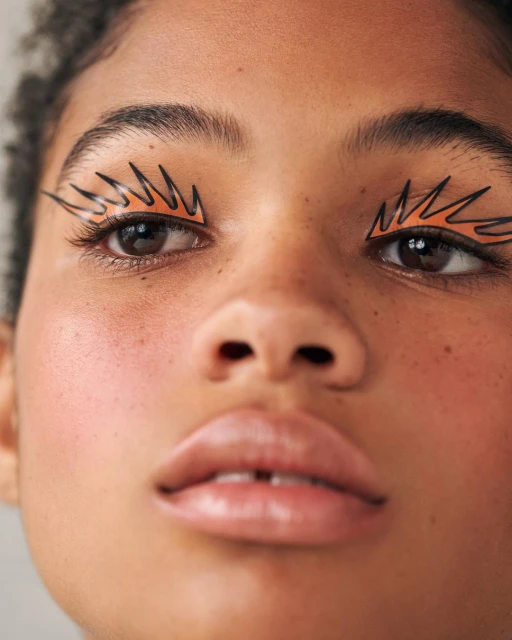 Close-up studio shot of a person with creative makeup featuring graphic orange eyeliner wings, neutral lip gloss, and fair skin.