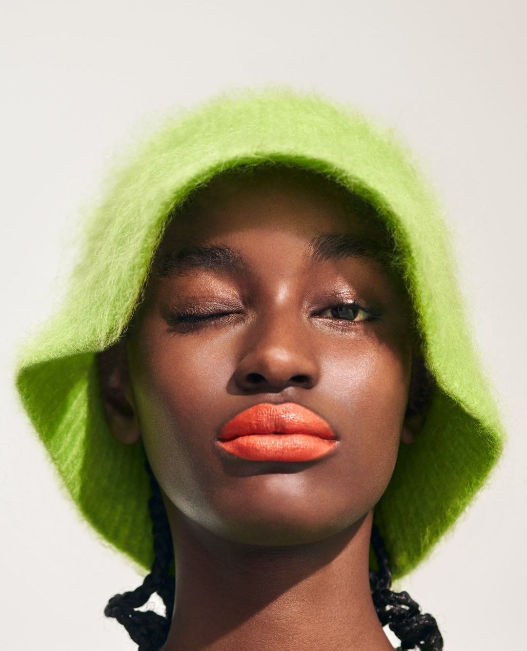 Close-up portrait of a winking Black woman wearing a fuzzy chartreuse bucket hat and vibrant coral lipstick, set against a plain background.