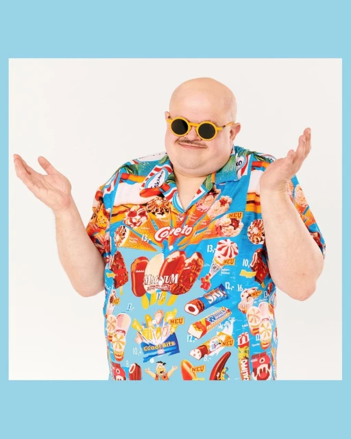 Bald adult man wearing round yellow sunglasses and a kitschy shirt printed with ice cream and candy, making a gesture with his hands.