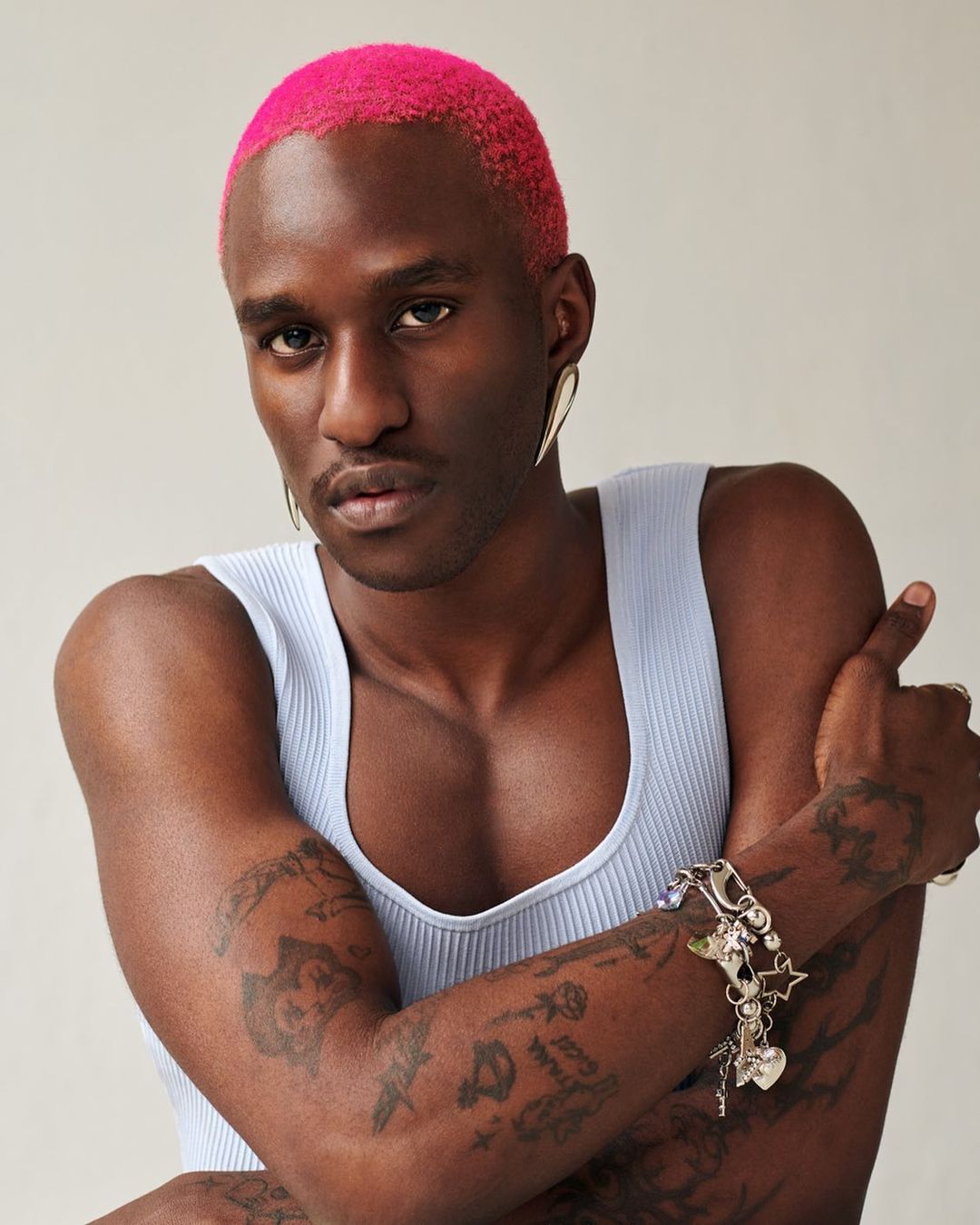 Portrait of a dark-skinned adult male with pink hair wearing a blue tank top, gold teardrop earrings, and a charm bracelet. Indoor setting.