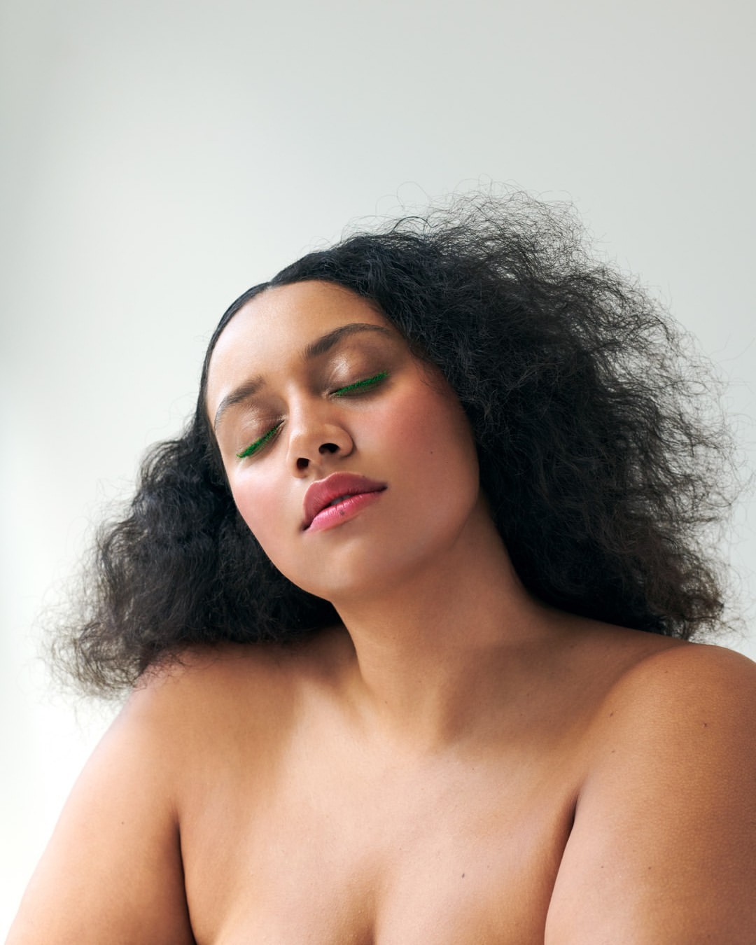Close-up studio portrait of a young woman with dark curly hair, wearing green eyeliner and pink lipstick, against a white backdrop.