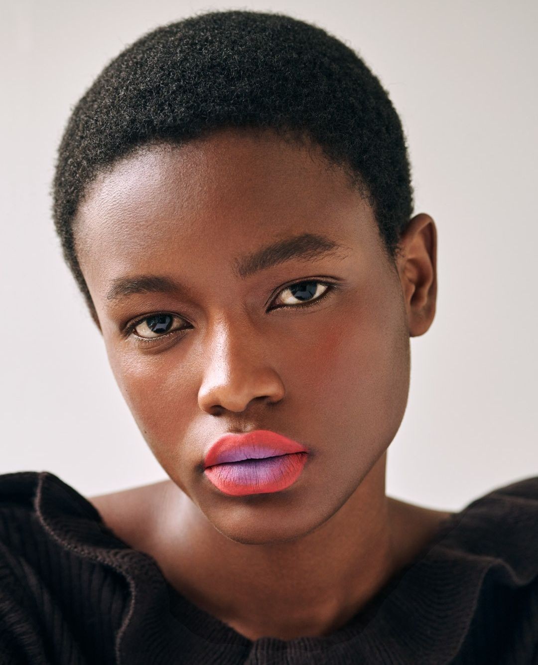 Close-up portrait of a young Black woman with short hair, wearing a black ruffled top and bold, two-toned ombre lipstick.