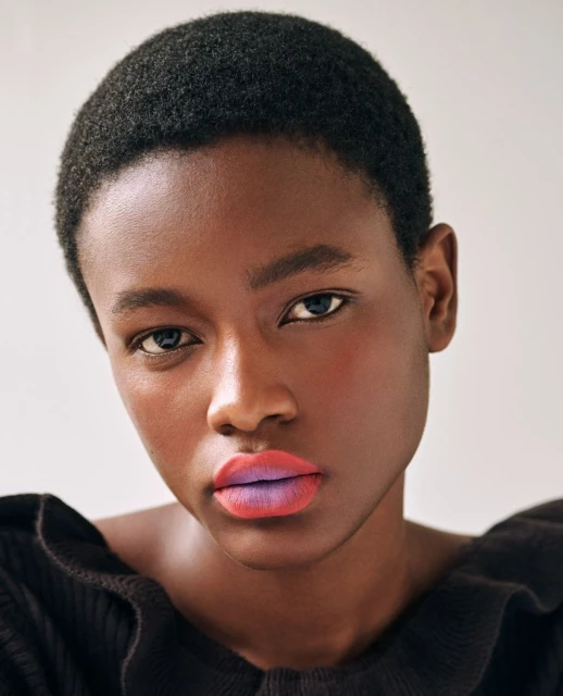 Close-up portrait of a young Black woman with short hair, wearing a black ruffled top and bold, two-toned ombre lipstick.