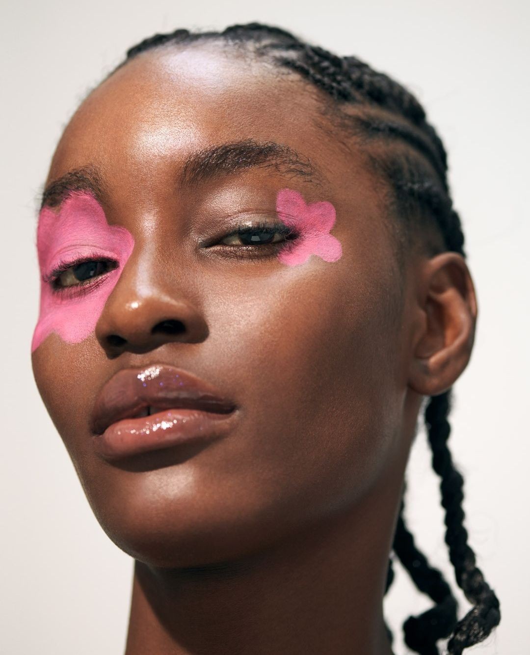 Close-up portrait of a black woman with braided hair and pink flower makeup art around her eyes against a white background.