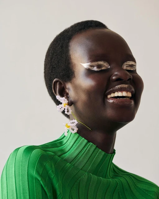 Close-up of a Black female model smiling with bold silver eye makeup, a bright green ribbed top, and beaded flower earrings.