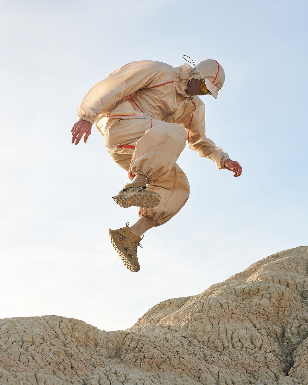 Person in beige hooded jumpsuit leaps over tan rocky hill against a pale blue sky, showcasing athletic footwear and stylish outerwear.