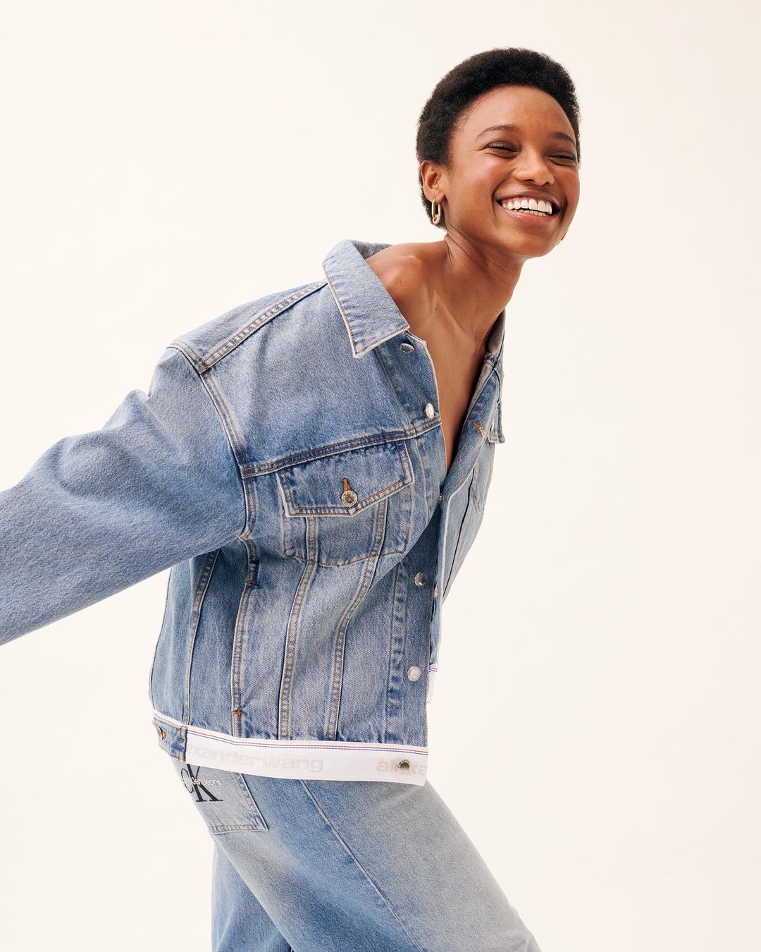 A smiling woman in a denim jacket and jeans poses in front of a white background in a fashion editorial shot with studio lighting.