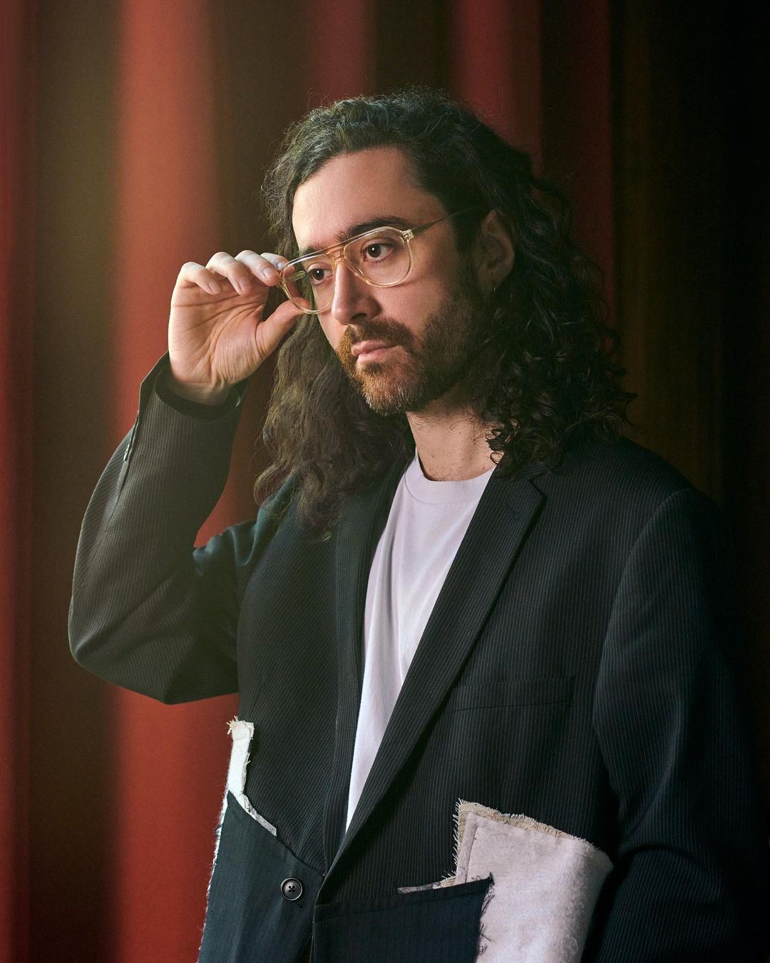Man with curly hair adjusts glasses in a medium studio shot, wearing a dark blazer, standing against a dark orange curtain.