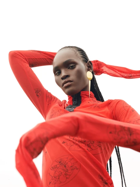 Woman with dark skin and braids poses in a red, long-sleeved top with a graphic print against a white backdrop wearing a gold earring.