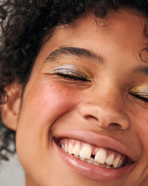 Close-up portrait of a smiling woman with curly hair and glittery gold and silver eye makeup against a plain background.